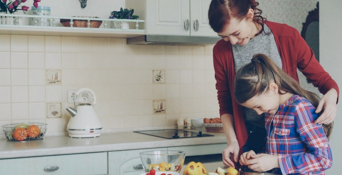 Young pretty mother teaching her cute daughter to cut vegetables properly. Little smiling girl cooking together with loving mom at home in modern kithcen