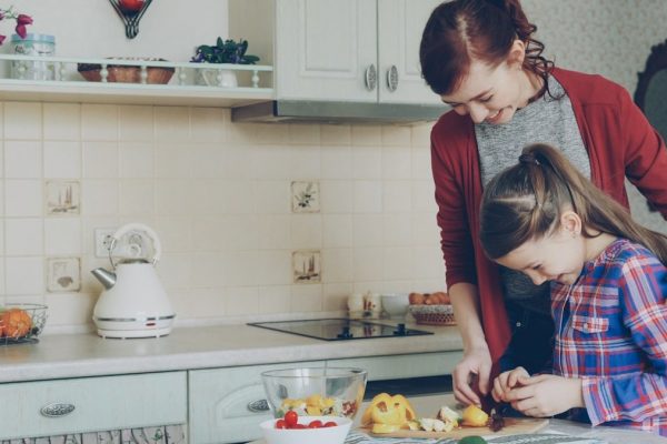 Young pretty mother teaching her cute daughter to cut vegetables properly. Little smiling girl cooking together with loving mom at home in modern kithcen